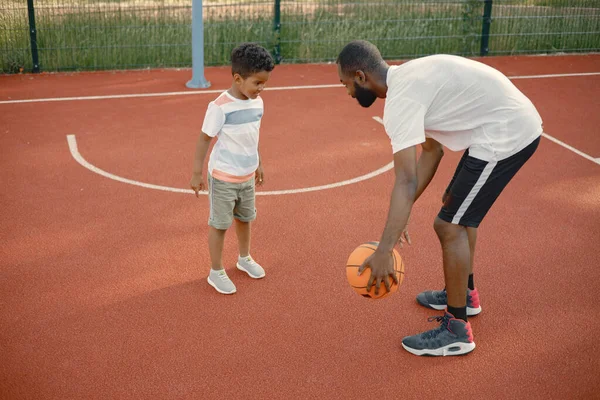 年轻的父亲和他的儿子在公园附近的篮球场打篮球 男人和男孩穿着白色的T恤 男人教他的儿子怎么玩 — 图库照片