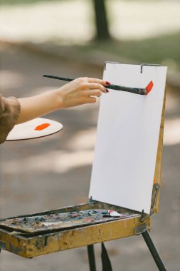 Açık havada boya üzerinde çalışan yetişkin kadın ressamın parçacık görüntüsü. Parkta resim yapan bir kadın. Beyaz kağıt üzerinde kırmızı boya darbeleri.