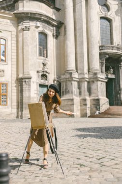Şehirdeki kadın ressam tablosu. Haki elbisesi ve siyah şapkası olan bir kadın. Kız, yazın kilisenin yakınındaki bir sehpada resim çiziyor..