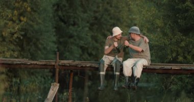 Baba ve oğul birlikte olta ve ağla balık tutuyorlar. Sabah vakti gölün ışığında ahşap iskelede oturuyorlar..
