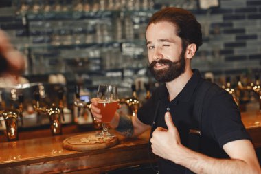 Attractive young man standing behind the bar. Bearded man drinking beer.
