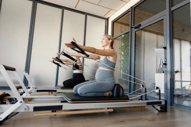 Özel bir ekipman üzerinde egzersiz yapan iki hamile kadın. Sarışın ve esmer kadınlar spor salonunda çalışıyor. Gri ve siyah spor ayakkabı giyen kadınlar..