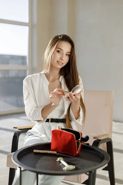 Young woman sitting on a chair and applying make up. Brunette woman sitting in a bright studio with big windows. Girl wearing white blouse and jeans.