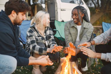 Çok ırklı arkadaşlar şehir dışında piknik yapıyorlar. Ateş yaktılar ve sıcak elleriyle oturdular. Avrupa, siyah, Pakistan ırkı. Minibüsle yolculuk..