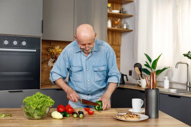 Yeşil salata, biber ve domateslerden yemek pişiren emekli son sınıf öğrencisi. Adam mutfağın içinde duruyor. Kot pantolon ve gömlek giyen adam..