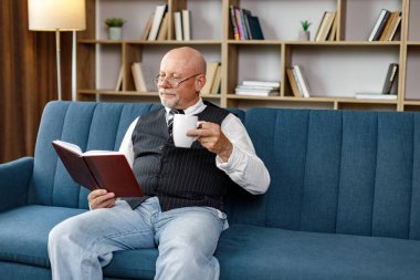 Kıdemli adam kanepede oturuyor ve akşamları evde kitap okuyor. Çay içen bir adam. Beyaz gömlek ve siyah yelek giyen adam..
