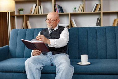Kıdemli adam kanepede oturuyor ve akşamları evde kitap okuyor. Çay içen bir adam. Beyaz gömlek ve siyah yelek giyen adam..
