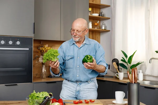 stock image Retired senior man cooking from green salat, pepper and tomatoes. Man standing in kitchen interior. Man wearing jeans and shirt.