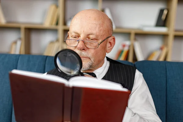 老年人晚上坐在沙发上看书 人使用放大镜 穿着白衬衫和黑色背心的男人 免版税图库照片