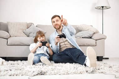 Sevgi dolu baba, Down sendromlu kızıyla evde birlikte oynuyor. Kız ve erkek kanepede oturup joystickleri tutuyorlar. Sakallı adam mavi gömlekli ve kız beyaz gömlekli..