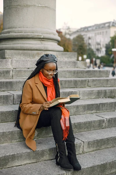 stock image Young black woman with a long locs hairstyles sitting on a stairs with a book. Woman wearing brown coat, orange scarf and black hat. Woman in glasses reading a book outside on autumn.