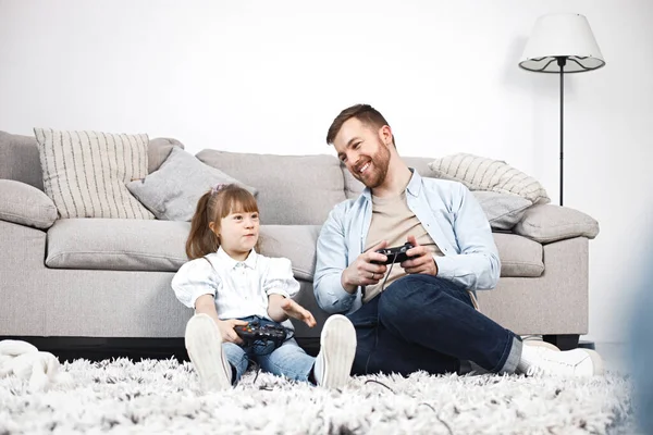 Loving father playing with his daughter with Down syndrome at home together. Man and girl sitting on a floor near sofa and holding joysticks. Bearded man wearing blue shirt and girl white shirt.