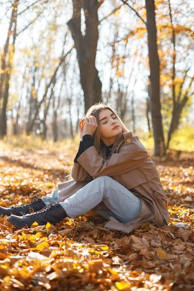秋の公園で写真を撮る美しい女性 若い女の子は黄色の葉の上に座っている ベージュのコートを着たブロンドの女性 — ストック写真