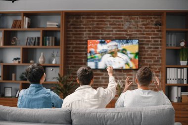 Bir futbol maçı izlemek için evde toplanan erkek arkadaşların arka görüntüsü. Erkekler büyük ekran televizyonun önünde kanepede oturuyor. Erkekler en sevdikleri takımı destekliyor ve maç hakkında yorum yapıyorlar.