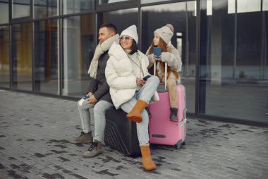 Family of three going on vacation. Man and woman sitting on a luggage and holding passports. Mother, father and daughter wearing warm clothes.