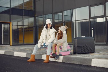 Brunette mother and her daughter going on vacation. Woman and girl sitting on a luggage and talking. Mother and daughter wearing warm clothes.