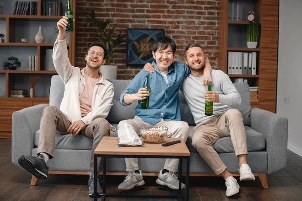 stock image Front view of male friends gathered at home to watch a football match. Men sitting on the couch in front of the big screen TV and eating snacks. Men actively support their favorite team and feeling