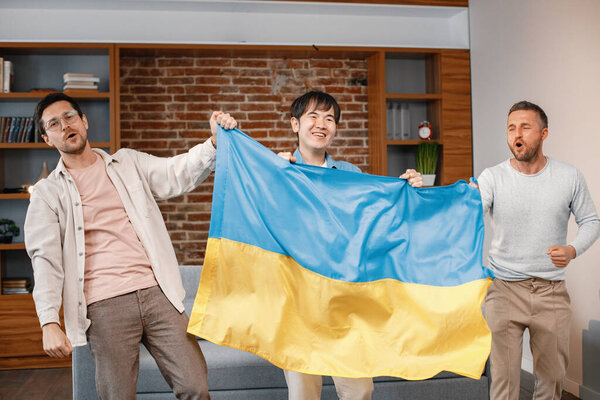 Male friends gathered at home to watch a football match. Men standing and holding a big flag of Ukraine in hands. Men actively support their favorite team and feeling satisfied.