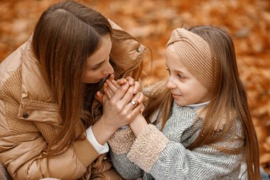 Young woman and little girl in autumn forest. Woman and her daughter sitting on a fallen leaves and holding hands. Girl wearing fashion grey jacket and woman brown one.