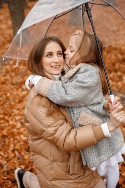 Young woman and little girl standing in autumn forest under transparent umbrella. Little girl hugging her mother. Girl wearing fashion grey jacket and woman brown one.