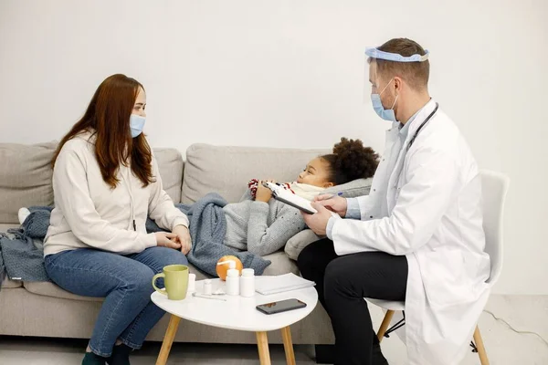 stock image Male doctor in white medical gown asking sick girl about her symptoms. Black girl has fever and overspread blanket. Different medicines are on table near sofa.