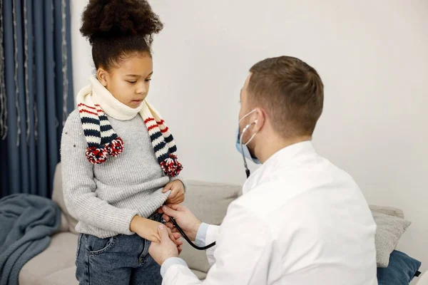 診察中の男性医師と病気の黒人の少女 スカーフをかぶってソファの近くに立っている少女 医者は膝をついて聴診器を使って — ストック写真