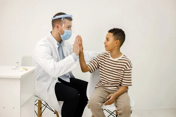 stock image Male doctor sitting at table and wearing face mask. Man wearing white medical gown while isolated on white backgroun. Man give a high five to a little boy.