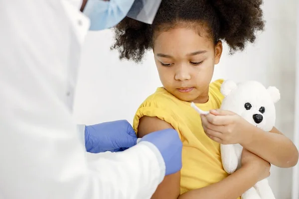 白衣を着た男性医師が予防接種後にバンドエイドを貼ります 女の子の顔の切り取られた写真 黄色のTシャツを着てクマのおもちゃを手に持っている女の子 ストックフォト