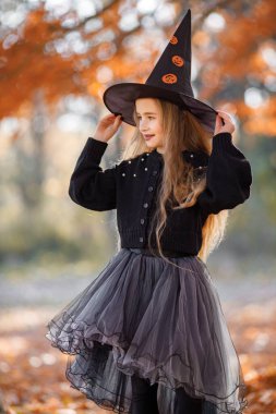 Cute happy little girl dressed in witch costume standing with over autumn forest background. Girl wearing dress and cone hat. Girl dressed for Halloween.