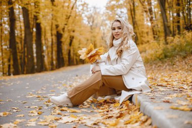 Young woman sitting on a curb in autumn forest. Blonde woman holding a yellow leaves. Girl wearing beige coat and brown trousers.