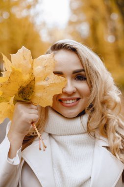 Young woman standing in autumn forest. Blonde woman holding a yellow leaves. Girl wearing white sweater.