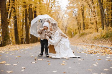 Sonbahar ormanında duran genç bir kadın. Sarışın kadın kızını öpüyor. Bej kazak ve beyaz şapka giyen ve elinde şeffaf bir şemsiye tutan bir kız..