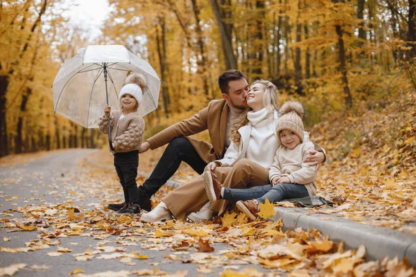 年轻的男人 女人和他们的孩子坐在秋天的森林外面 金发妈妈和金发爸爸拥抱在一起拿着一把透明的伞的小女孩 图库照片