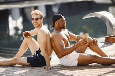 Beyaz ve siyah Amerikalı çocuklar yüzme havuzunun yanında dinleniyorlar. Beyaz ve siyah mayo ve güneş gözlüğü takan çocuklar. Kokteyl içen adamlar.