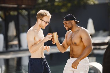 Beyaz ve siyah Amerikalı çocuklar yüzme havuzunun yanında dinleniyorlar. Beyaz ve siyah mayo ve güneş gözlüğü takan çocuklar. Erkekler kadehleri tokuşturuyor.