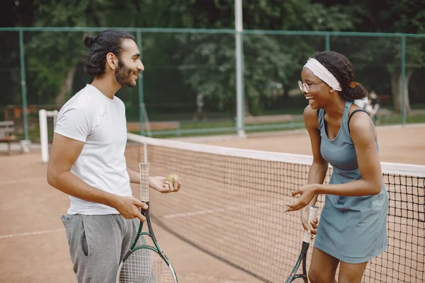 Indian man and black american woman standing on a tennis court. Woman wearing grey sport dress and man white t-shirt. Female and male tennis players talking.