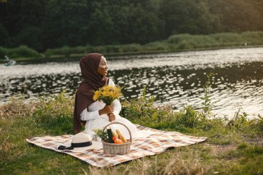 Tesettürlü Müslüman bir kız piknik yemeği ve bir buket çiçeğin yanında oturur. Nehir kenarındaki ekose piknik battaniyesinde kahverengi tesettürlü Müslüman bir kadının portresi. Beyaz giyen kız.