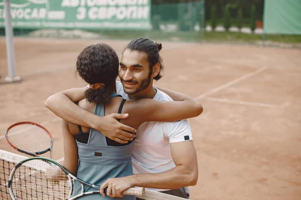Indian man and black american woman standing on a tennis court. Woman wearing grey sport dress and man white t-shirt. Female and male tennis players hugging after the match.