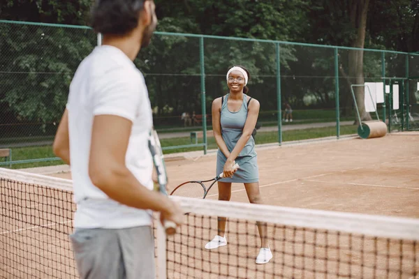 Focus on a woman. Indian man and black american woman playing tennis on a tennis court. Woman wearing grey sport dress and man white t-shirt.