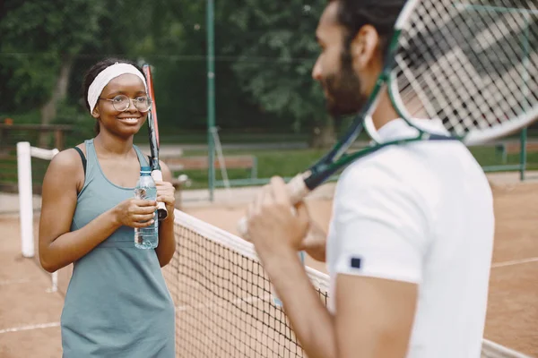 Indian man and black american woman standing on a tennis court. Woman wearing grey sport dress and man white t-shirt. Female and male tennis players talking.