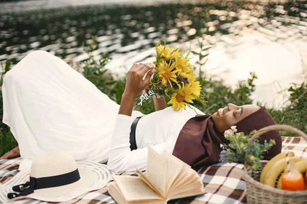 头戴头巾的穆斯林女孩手持一束鲜花躺在地上 在河边的格子布野餐毛毯上 有一位穆斯林女士头戴褐色头巾的画像 穿白衣的女孩 免版税图库照片