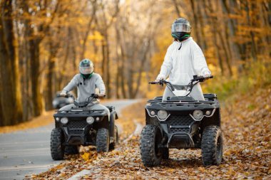 A young caucasian man and woman riding an ATV quad bikes in autumn forest. Two friends maneuvering off-road ATV. Couple wearing grey sportive costumes.