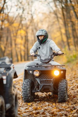 A young caucasian man in helmet riding an ATV quad bike in autumn forest. Boy maneuvering off-road ATV. Man wearing grey sportive costume.