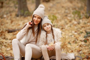 Young woman with little girl sitting on a fallen tree trunk in autumn forest. Brunette woman play with her daughter. Girl wearing beige sweater and mother wearing white clothes.