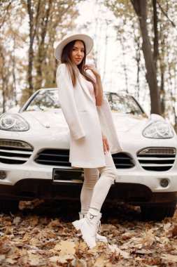 Portrait of woman in autumn forest, standing in front of her white car. Brunette woman posing for a photo. Woman wearing white hat, pink t-shirt and trousers.