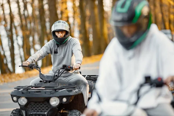 Focus on a man riding an ATV quad bikes in autumn forest. Two friends maneuvering off-road ATV. Couple wearing grey sportive costumes.