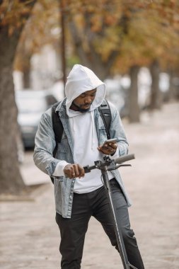 Elektrikli scooterlı siyah bir adam sokakta dikiliyor. Beyaz kapüşonlu ve siyah kotlu bir adam. Telefon kullanan siyah çantalı bir adam..