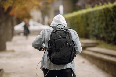 Adam siyah sırt çantası taşıyor ve sokakta yürüyor. Gri tişörtlü ve beyaz kapüşonlu bir adam. Sokaktaki bir adamın arka tarafı..