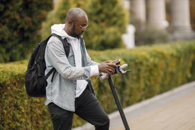 Elektrikli scooterlı siyah bir adam sokakta dikiliyor. Beyaz kapüşonlu ve siyah kotlu bir adam. Telefon kullanan siyah çantalı bir adam..