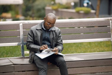Şehirdeki bir bankta oturan siyah bir adam. Güneş gözlüklü ve deri ceketli bir adam. Dışarıda çalışan bir iş adamı..